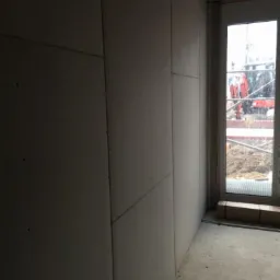 Wnętrze w trakcie prac wykończeniowych: ściany obłożone płytami gipsowo-kartonowymi, widoczne łączenia płyt, surowa podłoga, okno z widokiem na plac budowy i ciężki sprzęt.