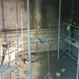 Ruthenia-Group Sp.z o.o - Murowanie ścian z szarej cegły wewnątrz budynku, widoczne rusztowania, podpory, oświetlenie robocze i pracownik w żółtej kamizelce na rusztowaniu.