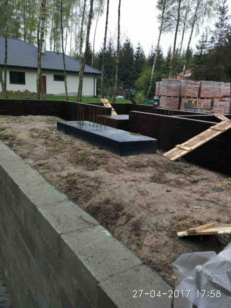 Fundamenty domu w trakcie budowy: betonowe ławy z wystającymi prętami zbrojeniowymi, częściowo zalane, otoczone szalunkiem i ziemią, w tle cegły i drzewa.