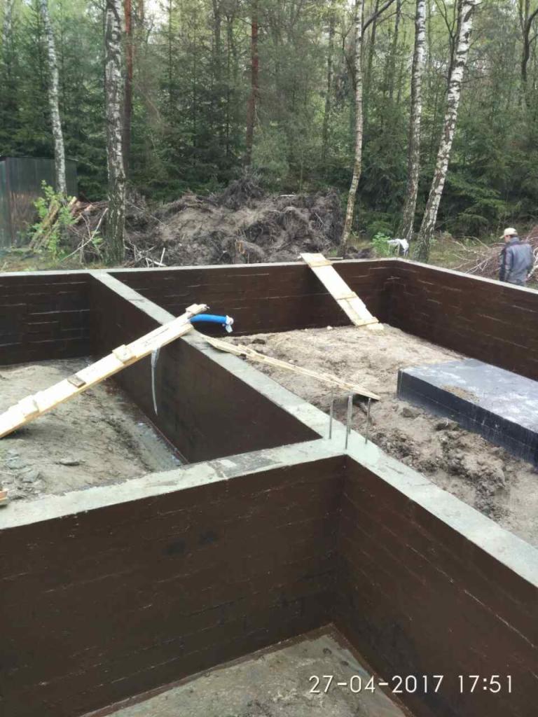 Wykop pod fundamenty domu z izolacją pionową ścian, widoczne wystające pręty zbrojeniowe oraz elementy szalunku, w tle las.