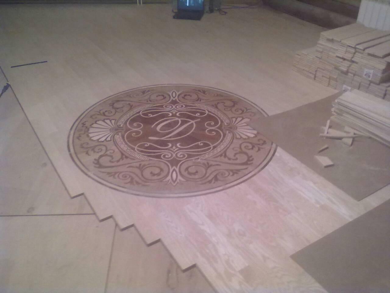 Układanie drewnianej podłogi z dekoracyjnym, okrągłym wzorem z literą 'D' w centrum, widoczne stosy desek podłogowych.