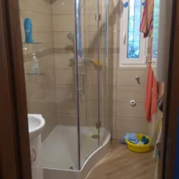 Ukończona łazienka z kabiną prysznicową z zaokrąglonym szkłem, beżowymi płytkami na ścianach i drewnopodobną podłogą, widok zza otwartych drzwi.