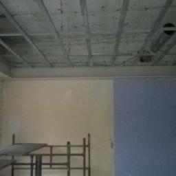 SzydlikPaweł - Wnętrze w trakcie prac wykończeniowych: widoczna konstrukcja sufitu podwieszanego, częściowo pomalowana ściana na dwa kolory oraz rusztowanie.