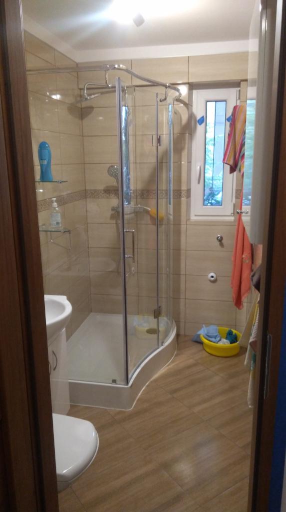 Ukończona łazienka z kabiną prysznicową z zaokrąglonym szkłem, beżowymi płytkami na ścianach i drewnopodobną podłogą, widok zza otwartych drzwi.