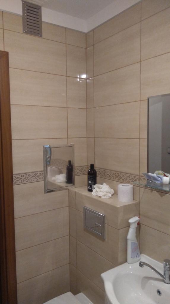 Beżowa łazienka z kwadratowymi płytkami, widoczna umywalka z baterią, zabudowana spłuczka, lustro w zabudowie, dekoracyjny pas płytek i kratka wentylacyjna pod sufitem.