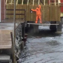 Pracownik w pomarańczowym kombinezonie, stojący na wąskiej platformie nad wodą, maluje drewnianą ścianę budynku w trakcie budowy, widoczne rusztowanie i wzburzona woda.