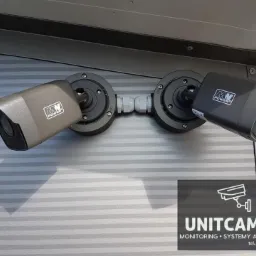 Dwie kamery monitoringu IP, szara i czarna, zamontowane na szarej, poziomo żłobkowanej ścianie pod kątem, połączone przewodem, z logo producenta MW Power.