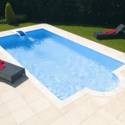 Prostokątny basen z jasną wodą otoczony beżowymi płytkami, z dwoma czarnymi leżakami z czerwonymi ręcznikami, na tle zielonego trawnika i żywopłotu.