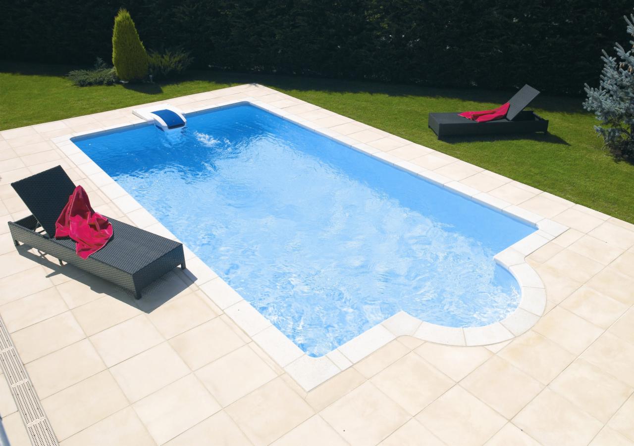 Prostokątny basen z jasną wodą otoczony beżowymi płytkami, z dwoma czarnymi leżakami z czerwonymi ręcznikami, na tle zielonego trawnika i żywopłotu.