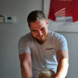 Uśmiechnięty mężczyzna w szarej koszulce z logo Salveo wykonuje masaż pleców pacjentce. W tle koszulka sportowa z napisem Polska.