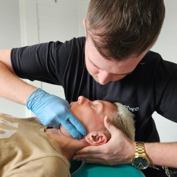 Salveo Medical Care Ruda Śląska Sp. z o.o - Terapeuta w niebieskich rękawiczkach wykonuje masaż twarzy pacjentowi leżącemu na turkusowym fotelu. Widoczny złoty zegarek na nadgarstku terapeuty.