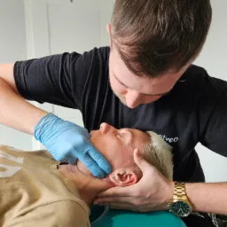 Terapeuta w niebieskich rękawiczkach wykonuje masaż twarzy pacjentowi leżącemu na turkusowym fotelu. Widoczny złoty zegarek na nadgarstku terapeuty.