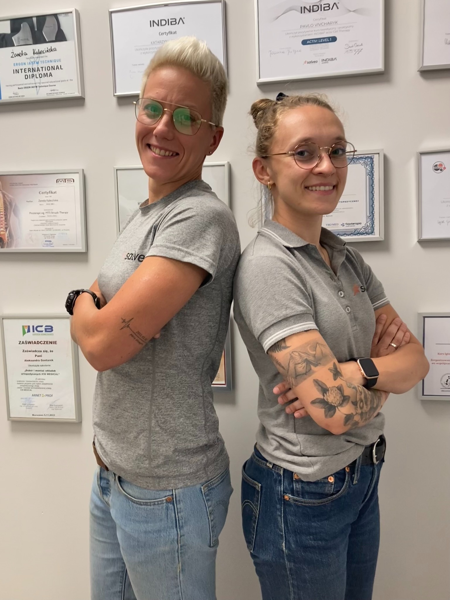 Dwie uśmiechnięte fizjoterapeutki, stojące tyłem do siebie na tle ściany z certyfikatami. Kobiety w szarych koszulkach polo i jeansach, z tatuażami na rękach.