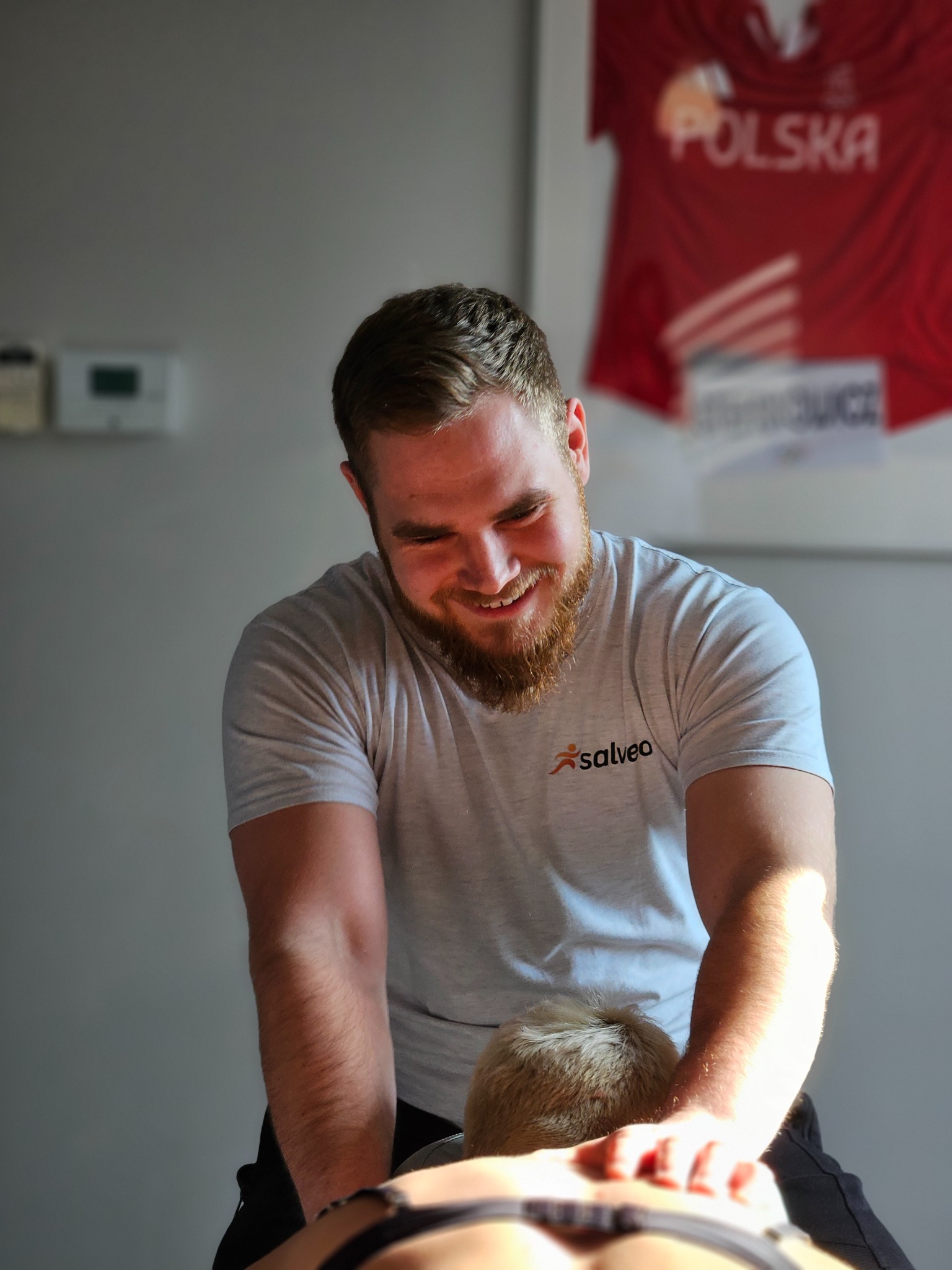 Uśmiechnięty mężczyzna w szarej koszulce z logo Salveo wykonuje masaż pleców pacjentce. W tle koszulka sportowa z napisem Polska.