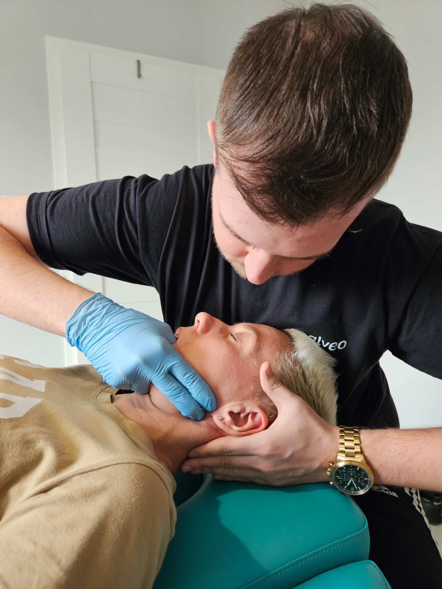 Terapeuta w niebieskich rękawiczkach wykonuje masaż twarzy pacjentowi leżącemu na turkusowym fotelu. Widoczny złoty zegarek na nadgarstku terapeuty.