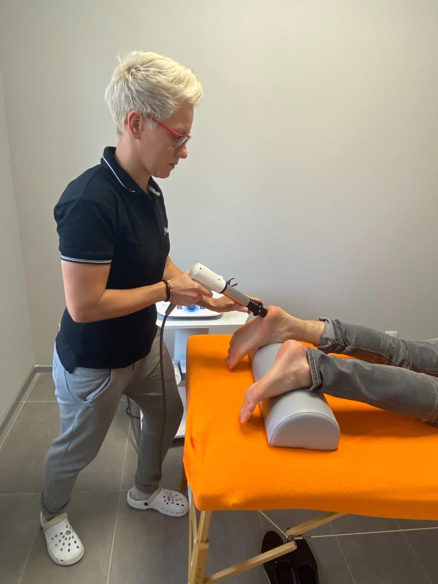 Kobieta w okularach wykonuje zabieg fizjoterapeutyczny na stopie pacjenta leżącego na pomarańczowym stole. Widoczny aparat do fizykoterapii.