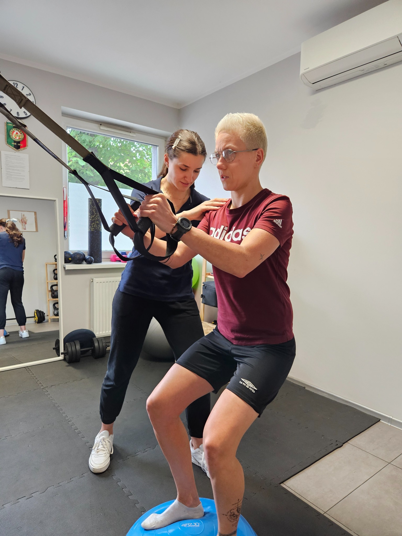 Osoba ćwiczy na bosu z pomocą trenera, używając taśm TRX. Widoczne lustro, zegar i sprzęt do ćwiczeń w tle. Jasne wnętrze gabinetu.