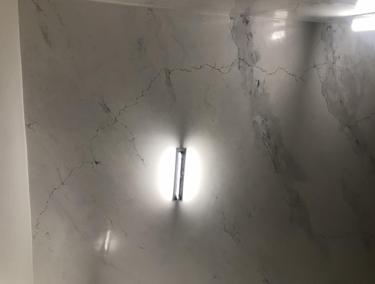 Ściana w jasnych odcieniach szarości z efektem spękań, oświetlona pionową lampą LED, imitująca marmur.