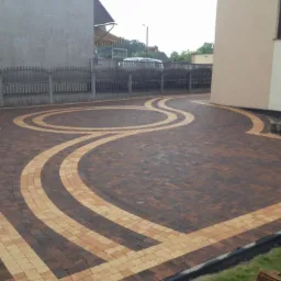 Ułożona dwukolorowa kostka brukowa w geometryczne wzory na podjeździe, z widocznym ogrodzeniem i fragmentami budynków w tle.