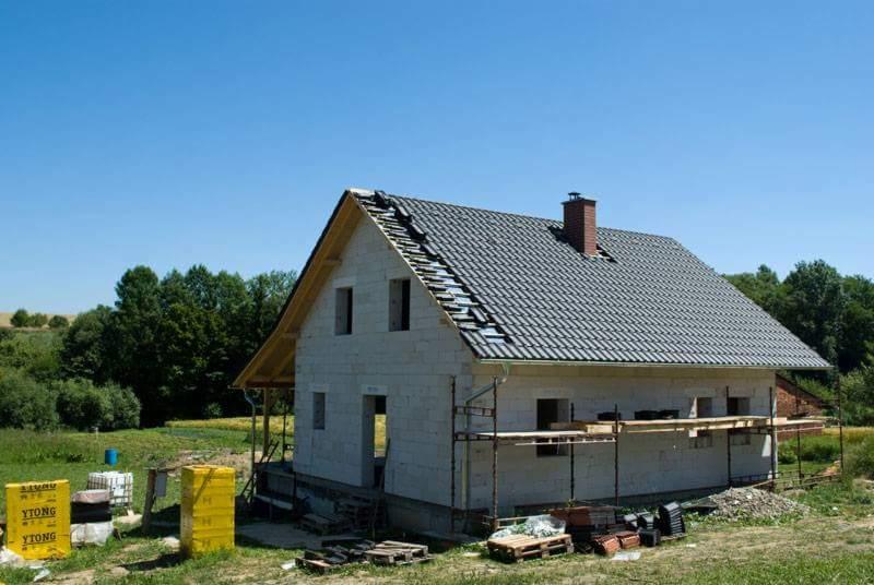 Dom w trakcie wymiany pokrycia dachowego, widoczne rusztowania, częściowo zdjęte stare dachówki i materiały budowlane YTONG na trawniku.