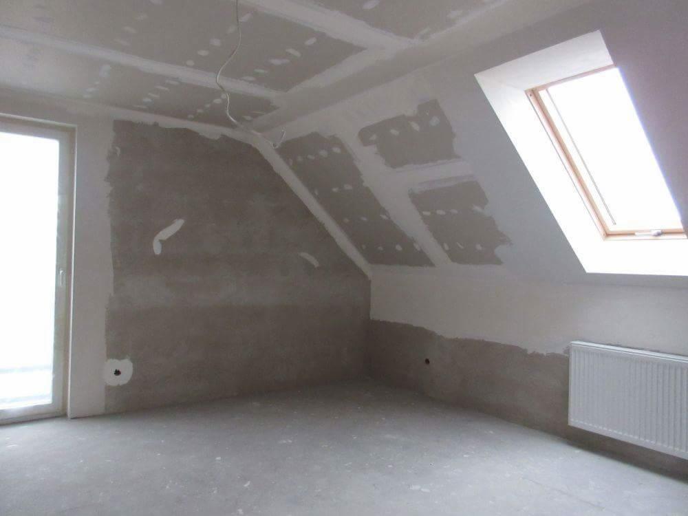 Pokój na poddaszu w trakcie wykańczania: ściany częściowo zagruntowane, widoczne płyty gipsowo-kartonowe na suficie, okno dachowe w drewnianej ramie, grzejnik na ścianie.