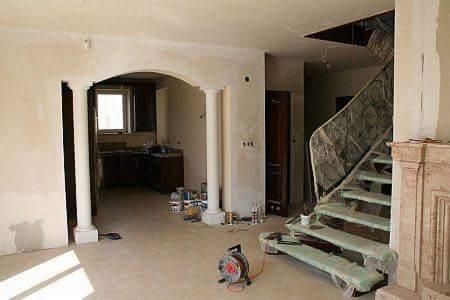 Wnętrze w trakcie remontu: surowe schody z metalową balustradą zabezpieczone folią, widok na kuchnię przez łuk z kolumnami, ściany w stanie surowym, narzędzia i materiały budowlane na podłodze.