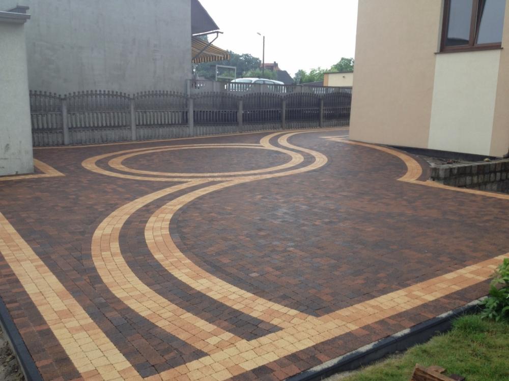Ułożona dwukolorowa kostka brukowa w geometryczne wzory na podjeździe, z widocznym ogrodzeniem i fragmentami budynków w tle.