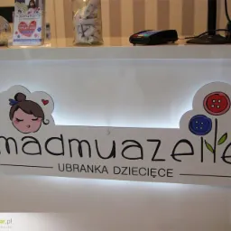 Podświetlany szyld frezowany z dibondu z grafiką przedstawiającą dziewczynkę i kwiaty z guzików, umieszczony na białej ladzie recepcyjnej w sklepie z ubrankami dziecięcymi 'madmuazene'.