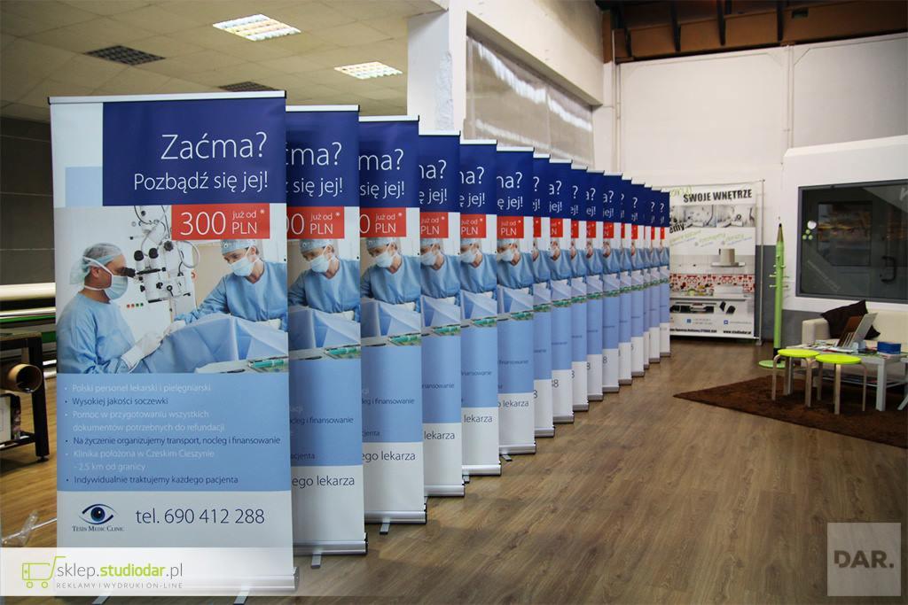 Rząd roll-upów z reklamą kliniki okulistycznej oferującej zabiegi usunięcia zaćmy, ustawionych na drewnianej podłodze w jasnym pomieszczeniu. Grafika na roll-upach przedstawia lekarza podczas...