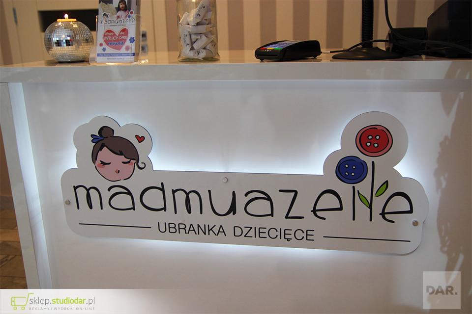 Podświetlany szyld frezowany z dibondu z grafiką przedstawiającą dziewczynkę i kwiaty z guzików, umieszczony na białej ladzie recepcyjnej w sklepie z ubrankami dziecięcymi 'madmuazene'.