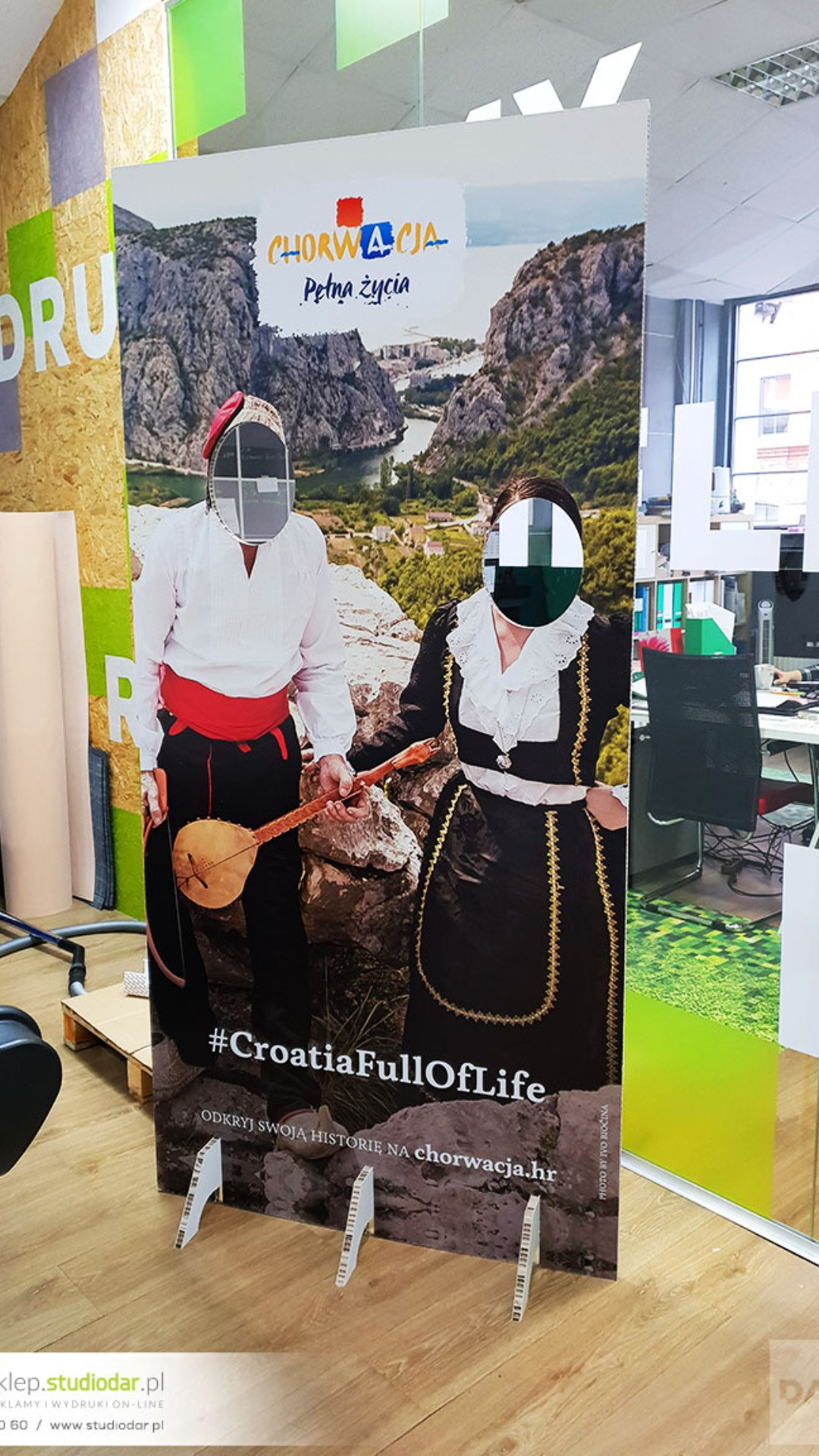 Duży, pionowy baner reklamowy z wyciętymi otworami na twarze, przedstawiający parę w strojach ludowych na tle chorwackiego krajobrazu, z hasłem #CroatiaFullOfLife i adresem strony internetowej.