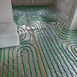 Zielone rury ogrzewania podłogowego ułożone na folii izolacyjnej z czerwoną siatką, widok z góry na surowy stan wykończenia wnętrza.