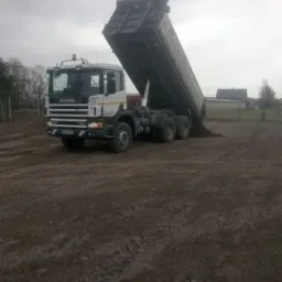 Ciężarówka Scania z podniesioną skrzynią ładunkową wysypuje ziemię na placu budowy, widoczne ślady opon na ziemi, pochmurne niebo w tle.