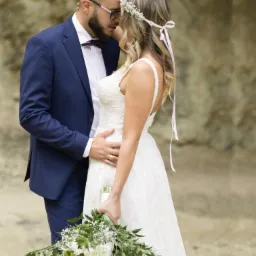 Para młoda: pan młody w granatowym garniturze i muszce, panna młoda w koronkowej sukni z wiankiem, delikatny bukiet ślubny, czuły gest dłoni na talii, naturalne tło skalne.