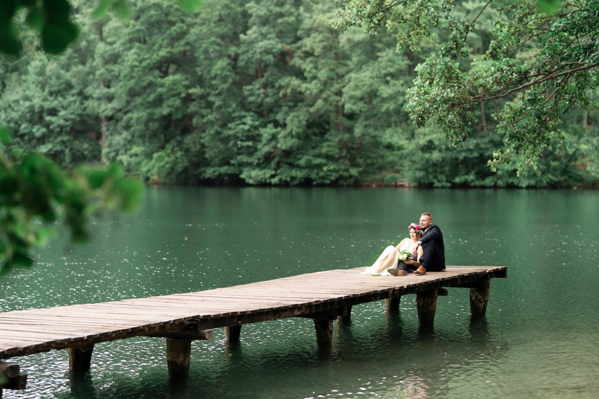 Para młoda siedzi na drewnianym pomoście nad jeziorem, w tle gęsty las, panna młoda w sukni i wianku, pan młody w garniturze.