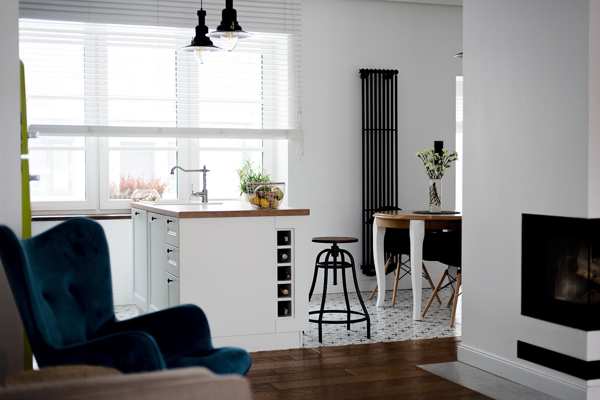 Jasne wnętrze kuchni z wyspą, stołkiem barowym i elementami dekoracyjnymi w Ksawerowie. Widoczny fragment salonu z turkusowym fotelem.