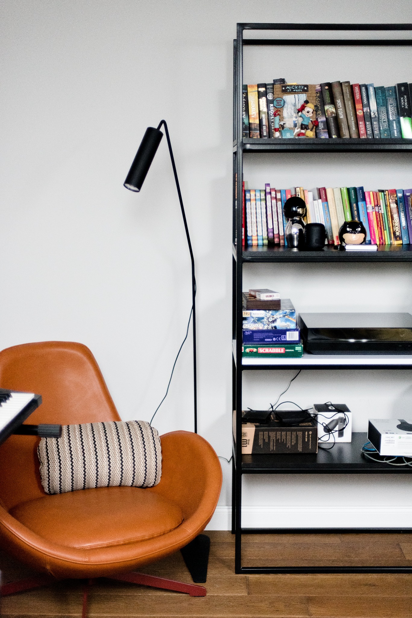 Pomarańczowy fotel z poduszką w paski, czarna lampa podłogowa i regał z książkami, grami planszowymi i dekoracjami w nowoczesnym pokoju.