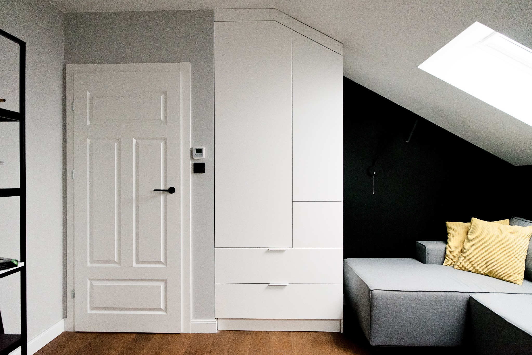 Biała zabudowa szafy wnękowej pod skosem na poddaszu, obok białe drzwi z czarną klamką, szara sofa z żółtymi poduszkami na tle czarnej ściany, oświetlenie górne ze świetlika.