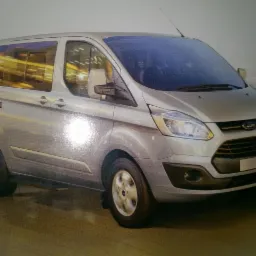 Srebrny Ford Transit Custom w wersji osobowej, widok z przodu i boku, z anteną na dachu i odbiciami w szybach.