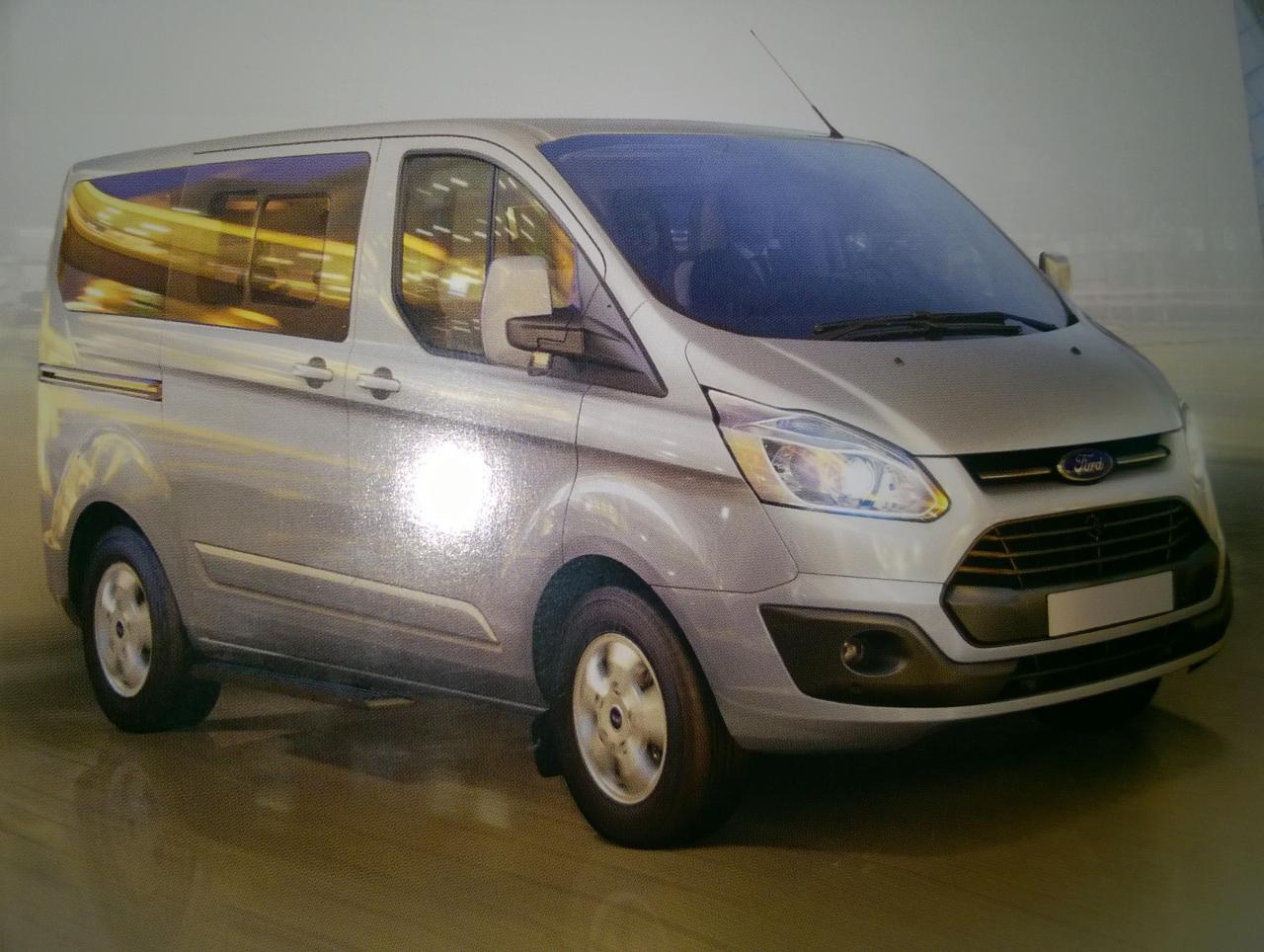 Srebrny Ford Transit Custom w wersji osobowej, widok z przodu i boku, z anteną na dachu i odbiciami w szybach.
