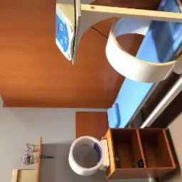 Aparat do rehabilitacji z niebieskim obiciem i białymi elementami konstrukcyjnymi, widoczny panel sterowania, drewniane wykończenie pomieszczenia, ujęcie z góry.