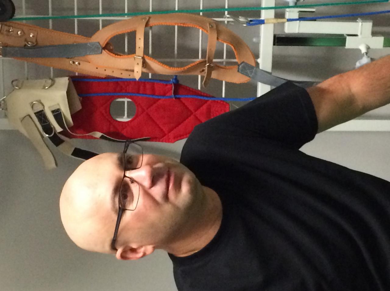 Fizjoterapeuta prezentuje sprzęt rehabilitacyjny: uprzęże i pasy do ćwiczeń w kolorach beżowym, czerwonym i białym, zawieszone na białej drabince.