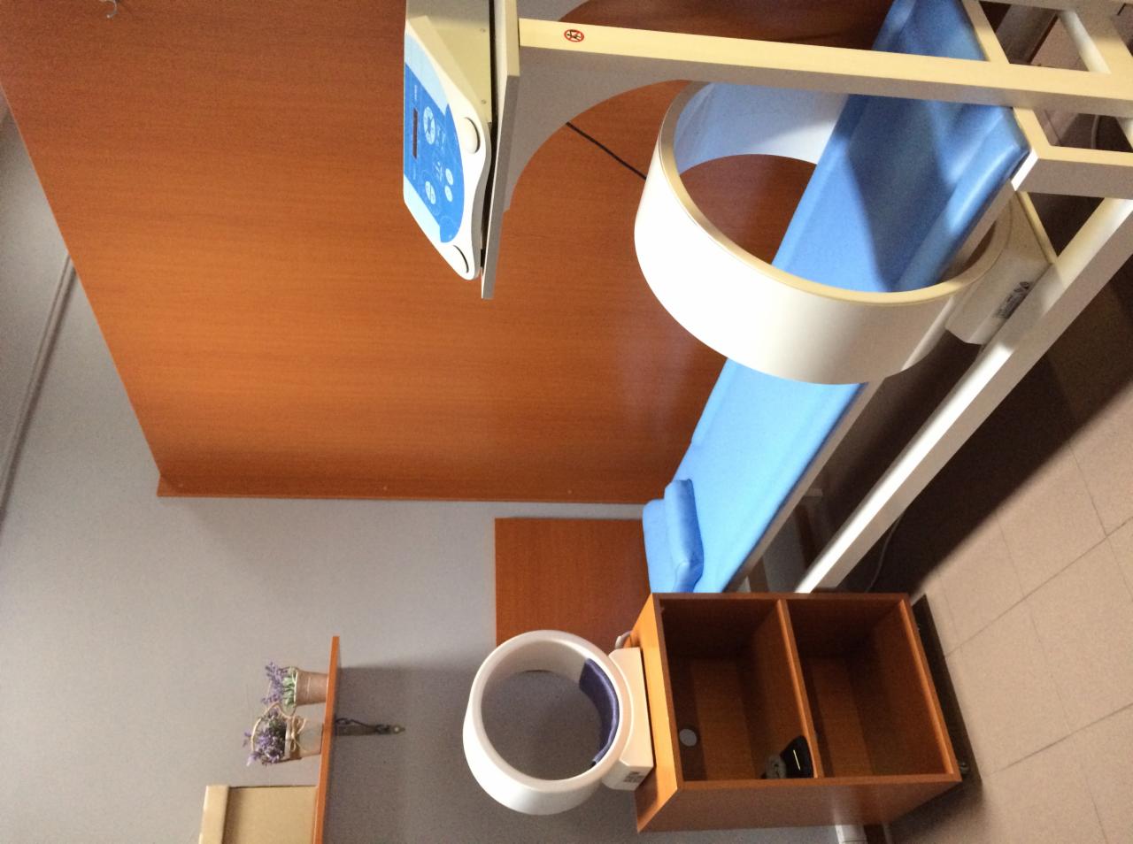 Aparat do rehabilitacji z niebieskim obiciem i białymi elementami konstrukcyjnymi, widoczny panel sterowania, drewniane wykończenie pomieszczenia, ujęcie z góry.