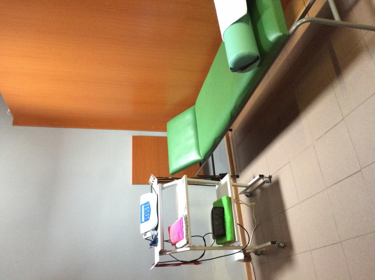 Zielony stół do zabiegów z regulowanym zagłówkiem i sprzętem do elektroterapii na wózku w pomieszczeniu z pomarańczowym sufitem.