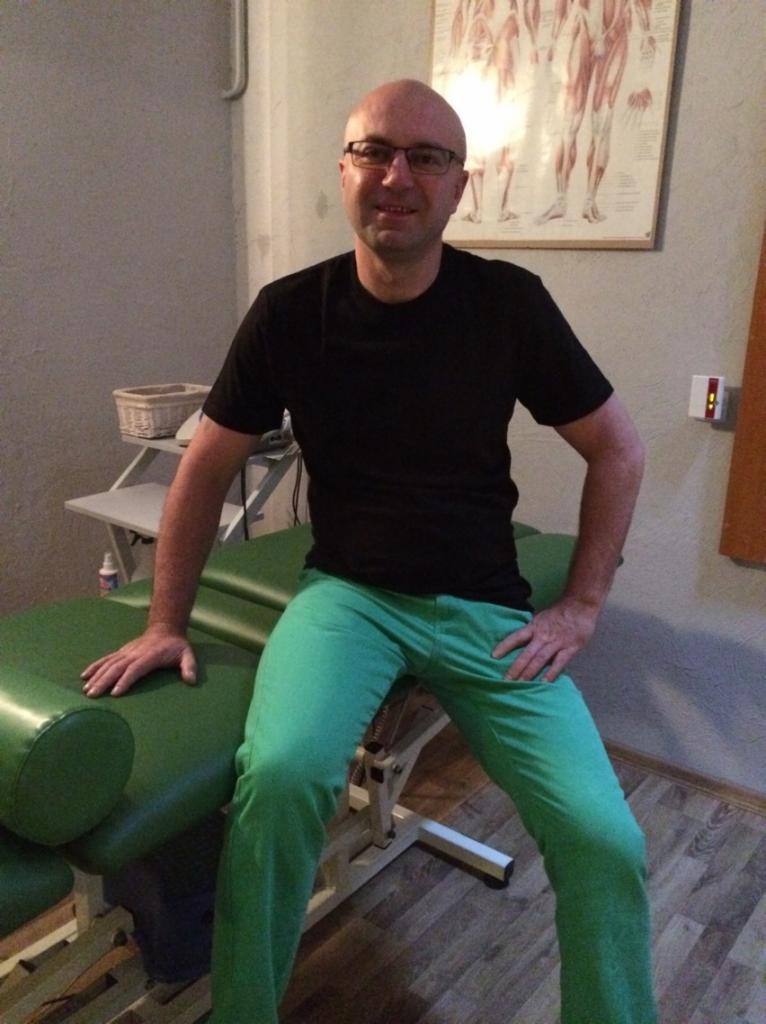 Mężczyzna w okularach siedzi na zielonym stole do zabiegów, w tle plakat z układem mięśniowym.