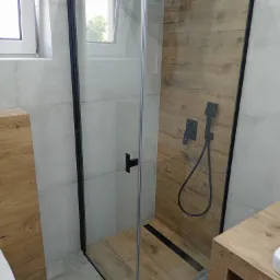 Nowoczesna kabina prysznicowa z czarnymi detalami i imitacją drewna na ścianach oraz podłodze, widok na okno z zielenią na zewnątrz.