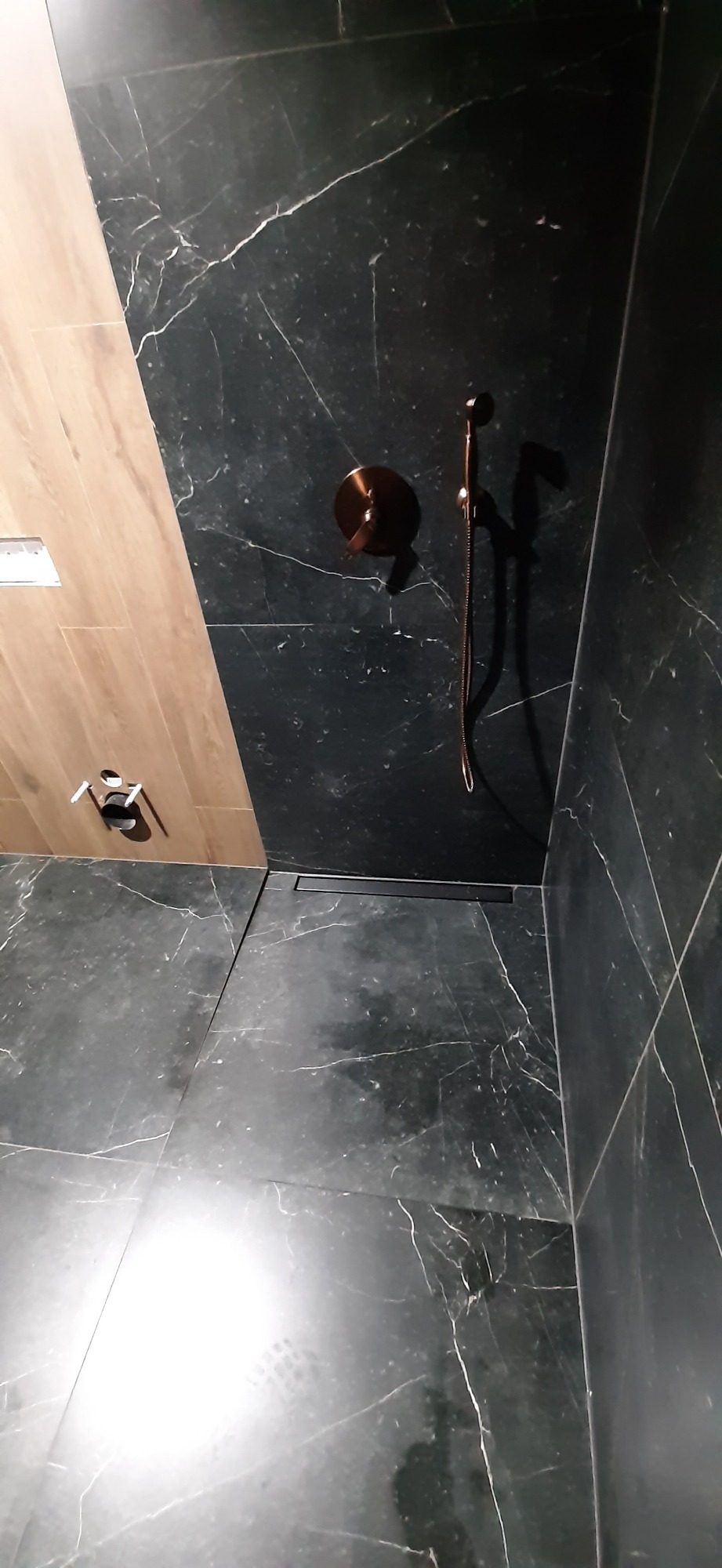 Nowoczesna łazienka z czarnymi marmurowymi płytkami, drewnopodobnymi panelami, odpływem liniowym i miedzianą armaturą natryskową.