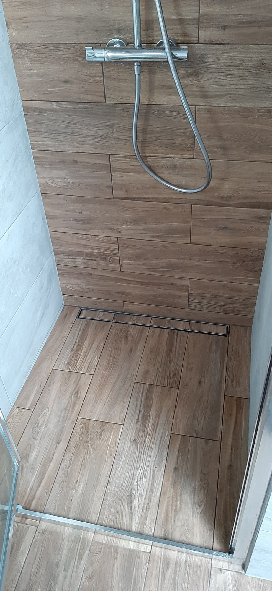 Kabina prysznicowa z odpływem liniowym, wykończona płytkami imitującymi drewno na ścianach i podłodze, widoczny chromowany zestaw natryskowy.