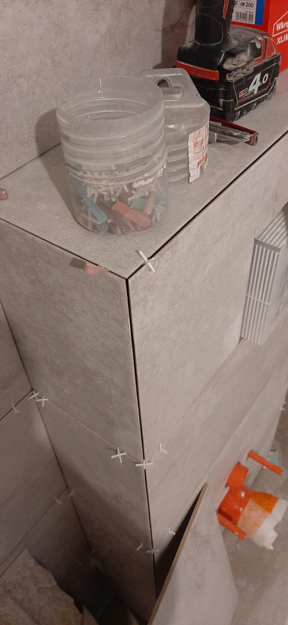 Fragment obudowy z płytek ceramicznych podczas prac wykończeniowych; widoczne krzyżyki dystansowe, pojemnik z klinami i wiertarko-wkrętarka akumulatorowa.