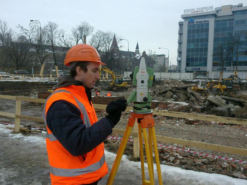 Mężczyzna w pomarańczowym kasku i kamizelce mierzy teren za pomocą niwelatora Leica na placu budowy z widocznymi wykopami i maszynami budowlanymi w tle.